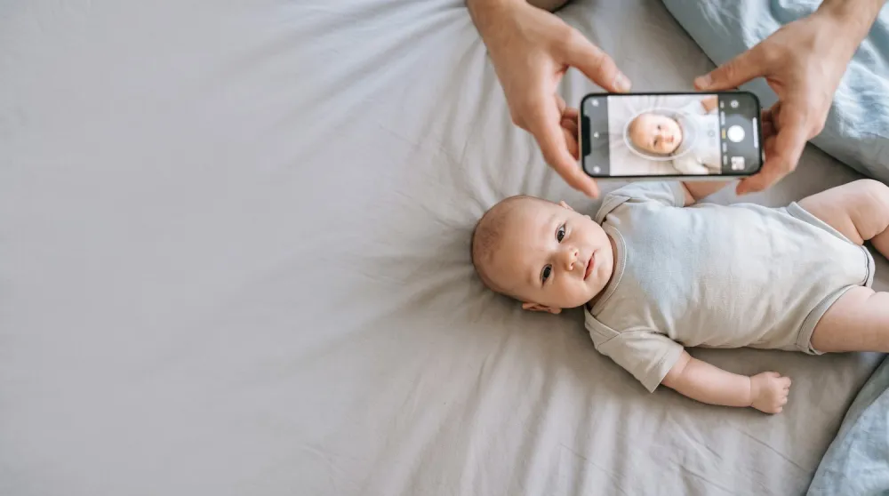 A baby lies on a light grey sheet and is photographed from above with a smartphone